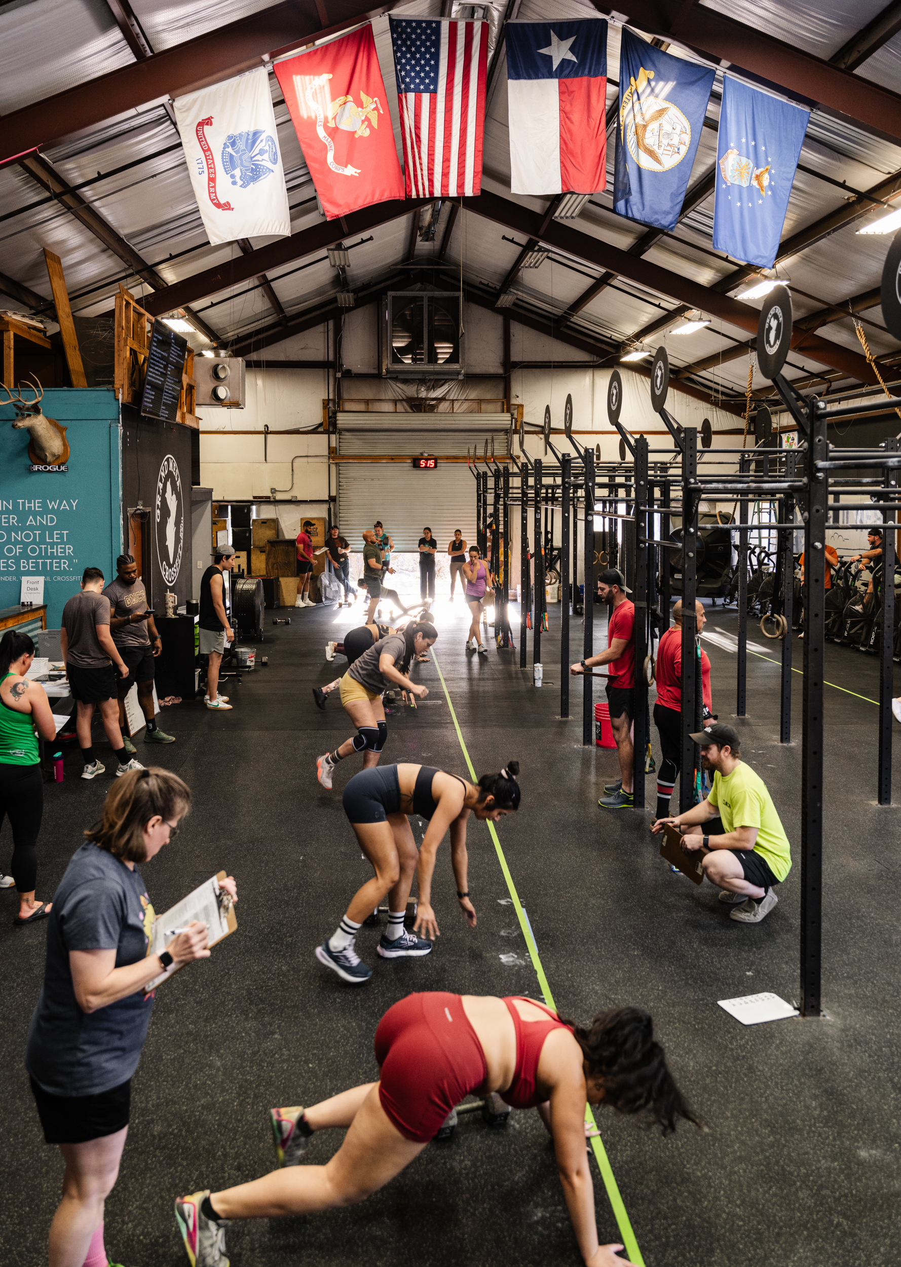 Group of athletes participating in a CrossFit workout inside a gym with rig stations, as coaches and spectators observe. The gym features multiple national and military flags hanging from the ceiling and motivational quotes on the wall.